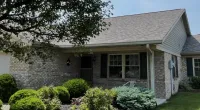Single-story brick house with gray roof, green shrubs, and a front porch with a white chair on a sunny day.