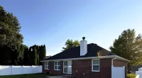 Single-story brick house with black roof, white fence, and green lawn under clear blue sky with Roof It Forward logo.