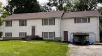White split-level house with brown roof and shutters, green lawn, gravel driveway, trees, and Roof It Forward logo.