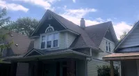 Two-story beige house with steep roof, white trim, brick pillars, and a porch under a partly cloudy sky.