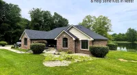Brick house with dark roof surrounded by green lawn, trees, and a pond under a cloudy sky.