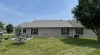 Brick single-story house with gray roof, green shutters, and a spacious lawn with trees under a clear sky.