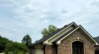 Brick house with a dark shingle roof on a cloudy day with greenery and a gravel driveway