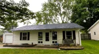 Single-story beige house with dark shutters, a front porch swing, and a green lawn surrounded by trees