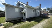Gray house with white trim and sloped roof under blue sky, driveway with red truck and Roof It Forward logo.