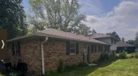 Single-story brick house with a large green lawn and tall trees under a partly cloudy blue sky.