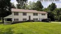 Two-story white house with brown roof and shutters on a green lawn under blue sky with Roof It Forward charity logo