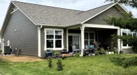 Single-story gray house with white trim, covered patio, lawn, potted plants, and outdoor seating under cloudy sky