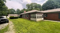 Brick ranch house with green lawn, gravel driveway, and trees under a cloudy sky