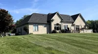 Large brick house with dark roof on expansive green lawn under bright blue sky