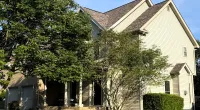 Two-story beige house with a sloped roof, surrounded by green trees and a well-maintained grassy lawn under blue sky.