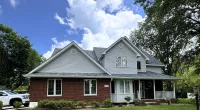 Two-story brick and siding house with a green lawn under a blue sky with clouds and a large tree.