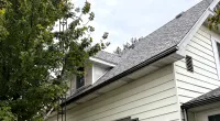 White house exterior with gray shingle roof, black shutters, tall tree, and overcast sky with Roof It Forward logo.