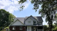 Suburban two-story house with gray roof, white siding, brick lower wall, surrounded by green trees and lawn under blue sky.