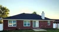 Single-story brick house with black shingle roof under clear sky, green lawn, and white fenced yard at dusk.