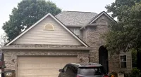 Suburban house with beige garage door, black SUV parked in driveway, and Roof It Forward charity logo.