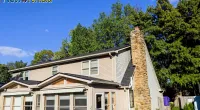 Two-story house with stone chimney, sunroom, and surrounding green trees under a clear blue sky.