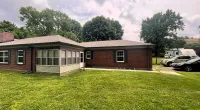 Red brick house with large green lawn, trees in the background, and cars parked near driveway under cloudy sky