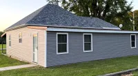 Gray house with new shingled roof and manicured lawn under clear sky near tall trees