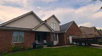 Modern brick and siding suburban house with a green lawn and a trailer parked in the driveway under a partly cloudy sky