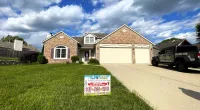 Brick house with a green lawn and driveway under a blue sky with white clouds, featuring a roofing company sign.