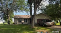 Suburban house with large trees, green lawn, and a car parked in the driveway on a sunny day.