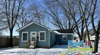 Blue house with snow-covered yard and bare trees under a clear winter sky with Roof It Forward logo.