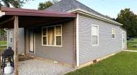 Grey house with a metal roof, covered gravel patio, and green grass lawn under a clear sky