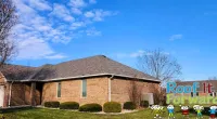 Brick house with a shingled roof under a blue sky, featuring Roof It Forward logo and cartoon children on the lawn.