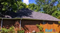 Brick house with a new shingled roof, surrounded by green trees, bushes and grass under a sunny sky.