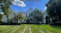 Large white house with green lawn and trees under a blue sky with clouds promoting Roof It Forward charity.