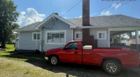 Red pickup truck parked beside a light gray house with chimney under a sunny blue sky with clouds.