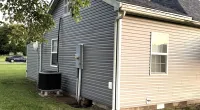 Side view of a house with two-tone vinyl siding, a grassy lawn, and an HVAC unit under a tree in daylight.
