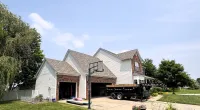 Suburban two-story house with an open garage, basketball hoop, trailer, and green lawn under a clear sky.