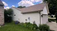 White detached garage with a sloped roof, green lawn, bushes, and a partly cloudy blue sky background.