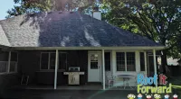 Backyard patio with covered roof, barbecue grill, white chairs, and table under shade from large trees.
