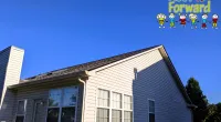 Suburban beige house with brown shingled roof under clear blue sky, Roof It Forward logo in top right corner.