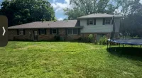 Spacious backyard with green grass, trampoline, and a two-story brick and siding house under a partly cloudy sky.