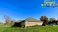 Single-story brick house with green lawn under clear blue sky and colorful Roof It Forward logo in corner