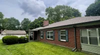 Single-story red brick house with green lawn, white windows, and surrounding trees under a cloudy sky.