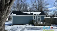 Snow-covered backyard with blue house, wooden deck, large tree, and clear blue sky in winter.