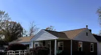 Single-story brick house with a brown roof, wheelchair ramp, bushes, and clear blue sky with Roof It Forward logo.
