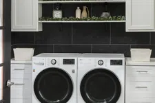 Thumbnail for Modern laundry room with white front-load washer and dryer, black tiled wall, white cabinets, and decorative shelves with plants.
