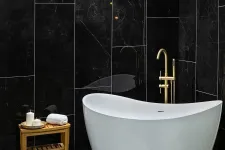 Thumbnail for Modern white freestanding bathtub with gold faucet against sleek black marble wall with wooden side table.