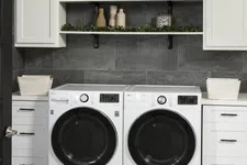 Thumbnail for Modern laundry room with white front-load washer and dryer, white cabinets, shelves with decor, and gray tiled wall.