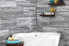 Thumbnail for Modern white freestanding bathtub with gray stone tile walls and decorative shelves in bathroom corner.