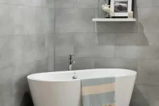 Thumbnail for Modern white freestanding bathtub with a striped towel in a gray tiled bathroom with a floating shelf above.