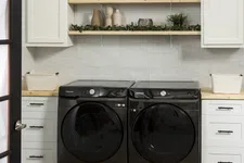 Thumbnail for Modern laundry room with black Samsung washer and dryer, white cabinets, wooden shelves, and decorative greenery.