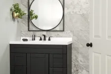 Thumbnail for Modern black bathroom vanity with white countertop, round mirror, gray tiled wall, and hanging plant in gold pot.