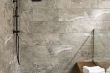 Thumbnail for Modern bathroom shower area with beige stone tiles, black rainfall showerhead, and wooden bench with white towels.